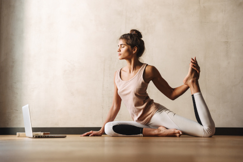 A woman does yoga on a mat in front of a laptop in a smaller room. 